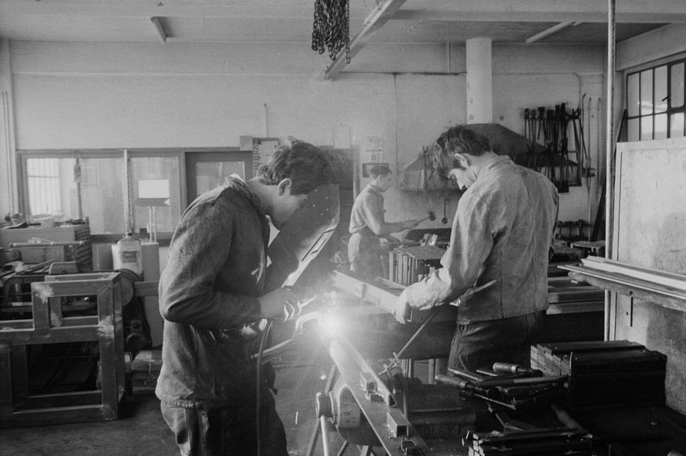 Tre apprendisti lavorano in un'officina per la lavorazione dei metalli. In primo piano si svolgono operazioni di saldatura e più indietro si lavora il metallo con gli attrezzi.