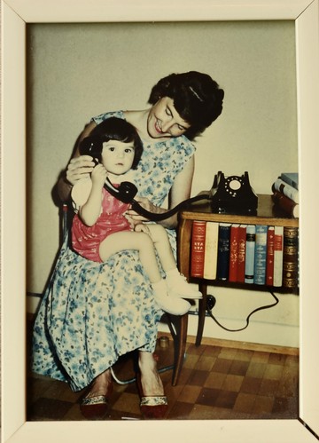Jasmin Schweizer*, di circa 2 anni, è seduta sulle ginocchia della madre e tiene in mano un grande ricevitore telefonico nero. La madre si china verso di lei e sorride. Su una piccola libreria alla sua destra si trova un telefono nero con un disco di selezione bianco.
