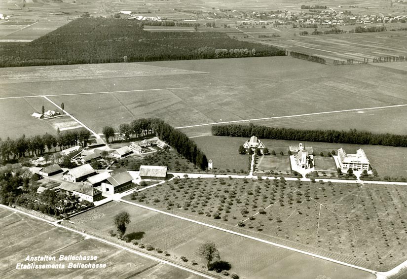 La veduta aerea mostra la caserma degli internati sulla sinistra, le officine e gli edifici agricoli, circondati da alberi. A destra si trovano la villa del direttore, la chiesa e l'ala della prigione.